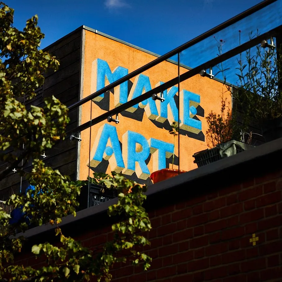 A colourful mural reading “MAKE ART” in large blue letters on the side of an orange building, framed by greenery and a glass balcony.